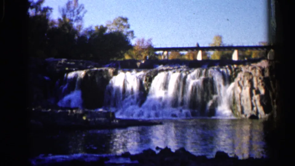 1960 8mm view of Sioux Falls, South Dakota, showing water cascading over rock ledges with a bridge in the background.