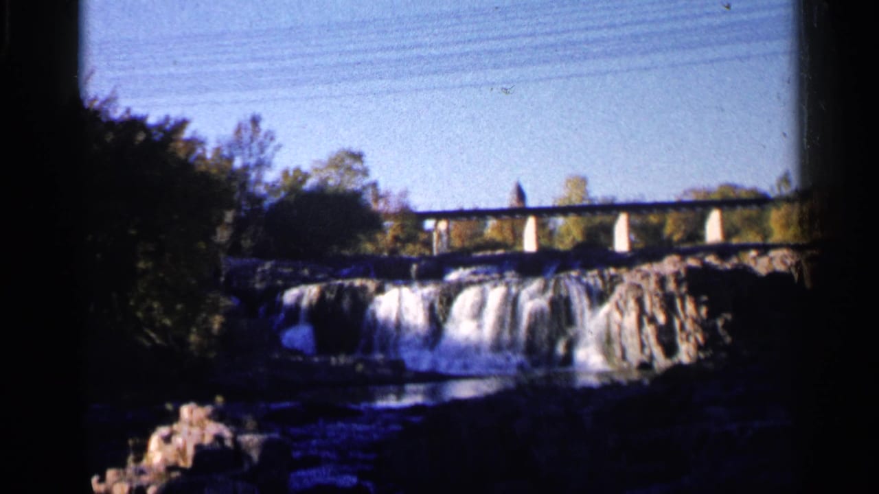 1960 8mm frame from Sioux Falls showing a bridge crossing above the waterfall, with trees and shadowed rocks around the river.
