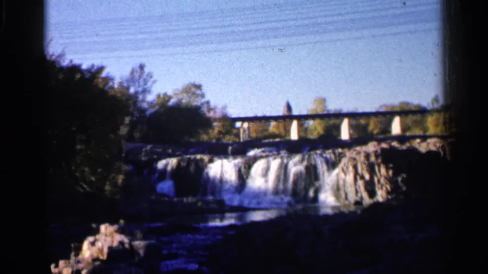1960 8mm frame from Sioux Falls showing a bridge crossing above the waterfall, with trees and shadowed rocks around the river.
