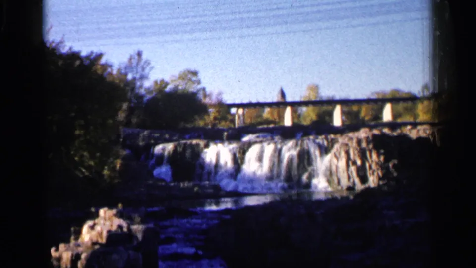 Visitor-shot 1960 8mm view of Sioux Falls, with cascading water, a bridge overhead, trees, and foreground rocks.