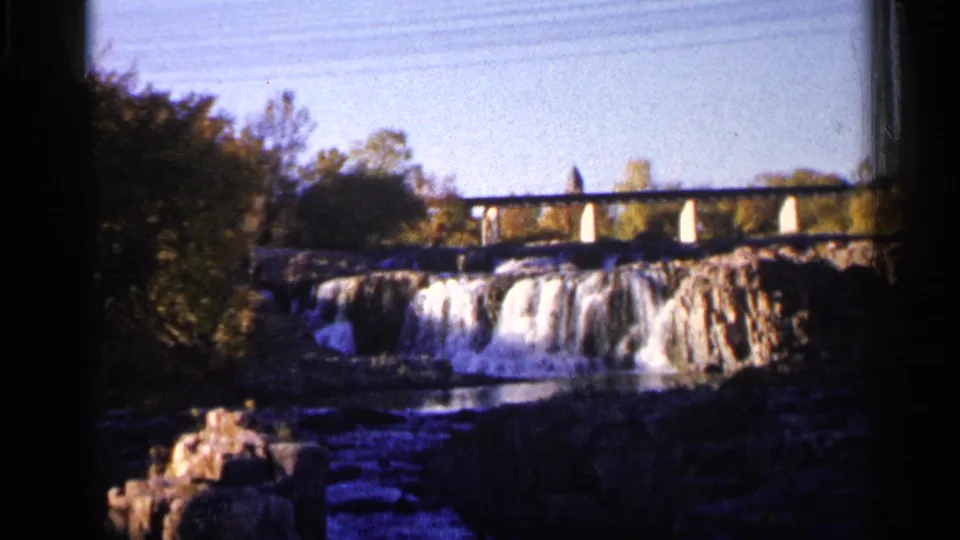1960 8mm Sioux Falls frame showing the waterfall, bridge, trees, and rocky river foreground in a steady home-movie view.