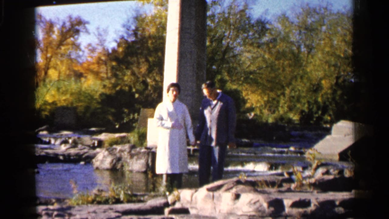 Man and woman standing on rocks beside a shallow river and small cascade.