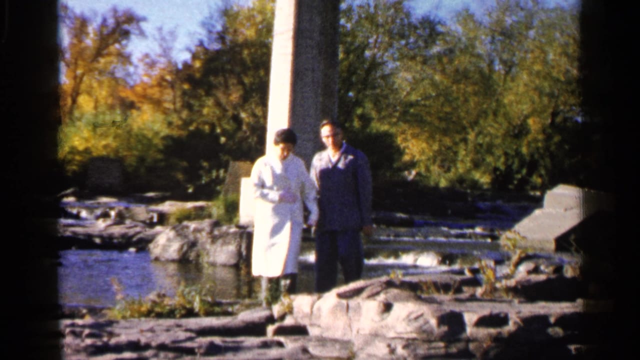 Two adults walking together on rocks near a river and concrete support.