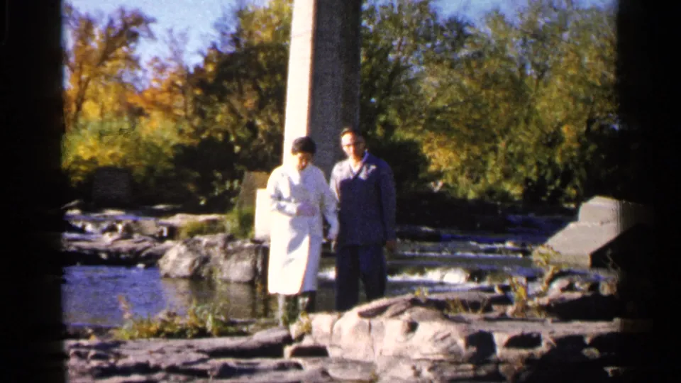 The couple walks along rocks by the river in Sioux Falls, with autumn foliage and a bridge-like concrete column behind them.