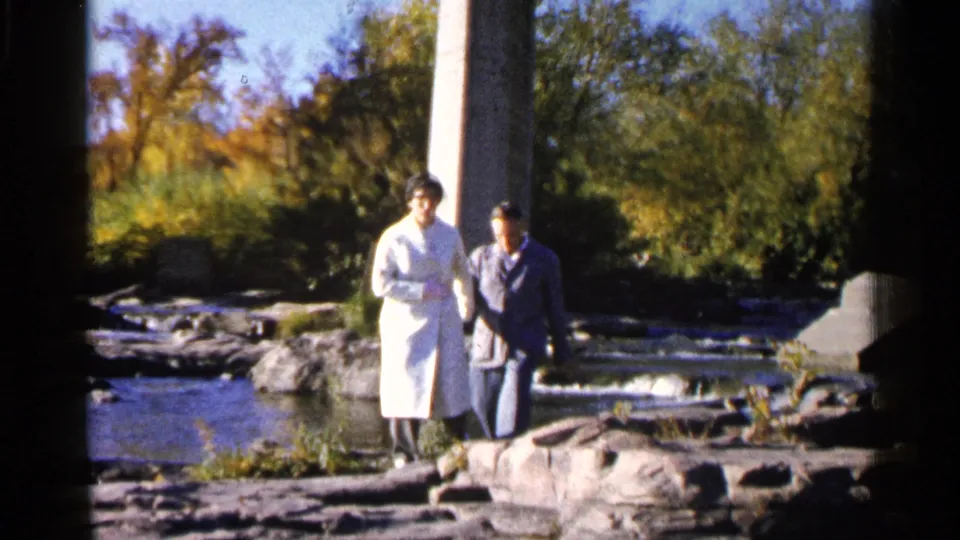 A woman in a long white coat and a man in dark clothes step over river rocks in 1960 Sioux Falls home-movie footage.