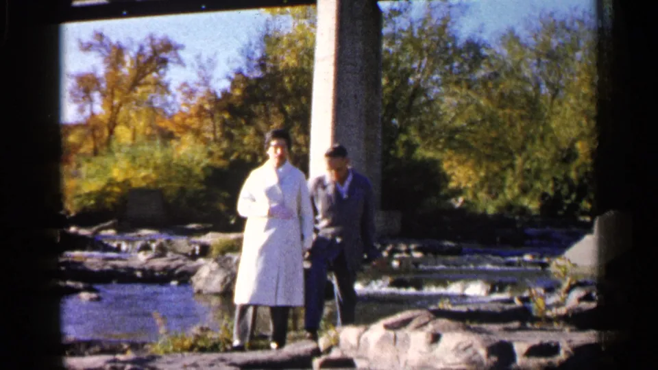 The woman and man stand near the edge of a rocky riverbed, with shallow water, autumn trees, and a concrete support visible.