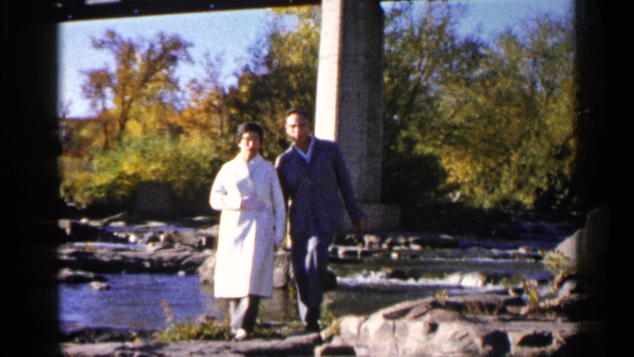 Couple walking beside a rocky stream under a bridge on a sunny autumn day.