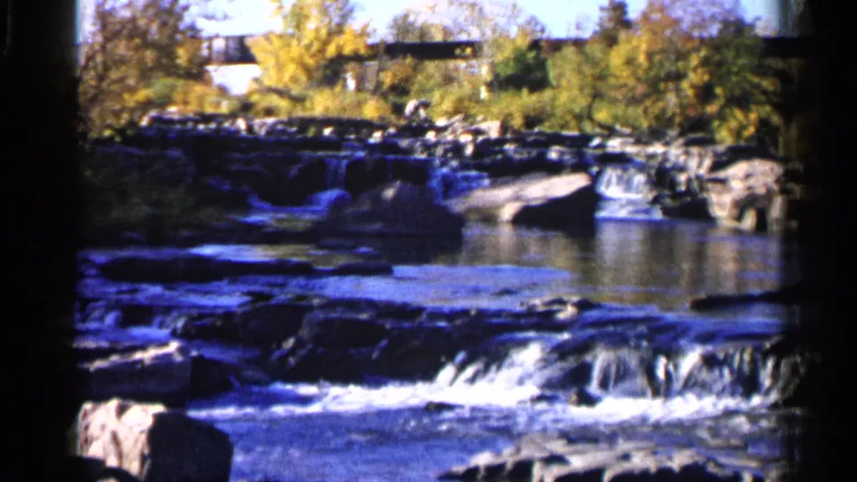 A Sioux Falls stream flows over low rocks with autumn foliage and bridge structures visible in 1960s 8mm home-movie footage.