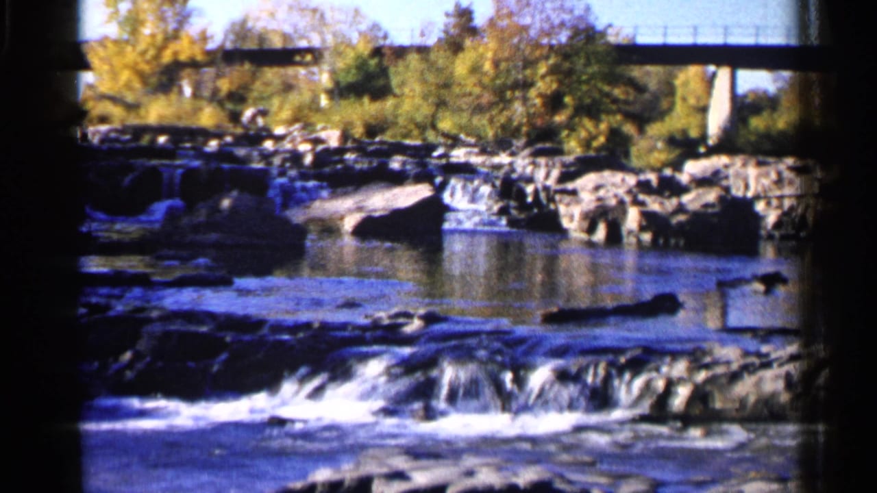 Rocky river rapids with a concrete bridge and autumn trees in the background.