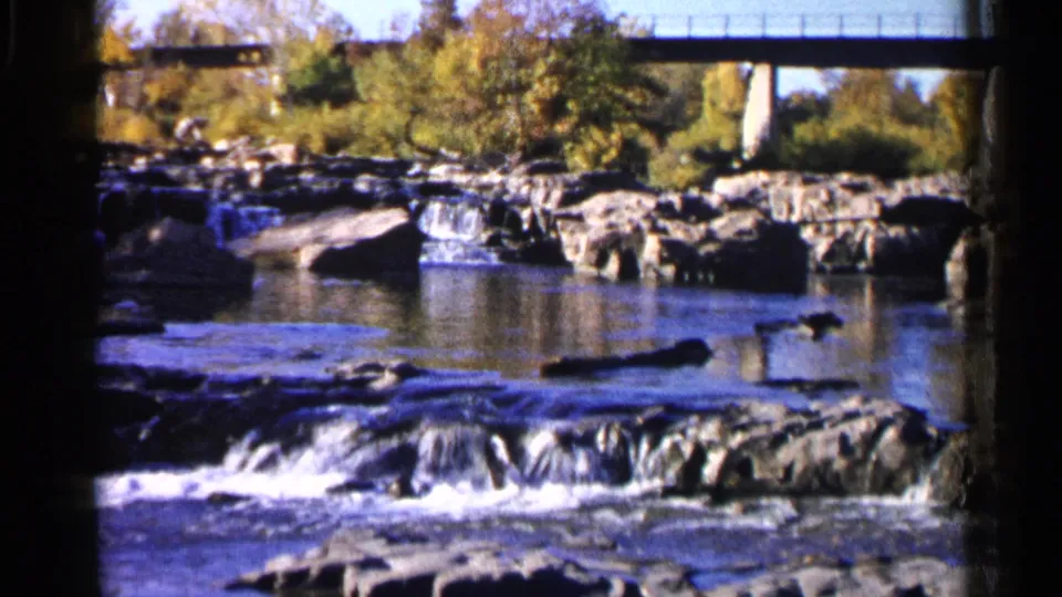 1960 Sioux Falls frame of shallow rapids, exposed rocks, a still pool, autumn foliage, and a concrete bridge in the background.