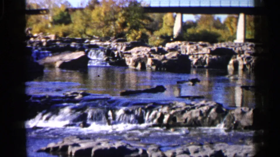1960 Sioux Falls home-movie view of rocky river ledges, white water, calm pools, autumn trees, and bridge supports.