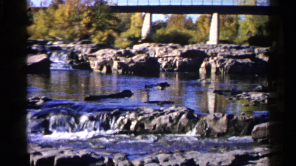 1960 Sioux Falls frame showing a rocky channel, rippling water, white rapids, autumn trees, and the concrete bridge above the river.