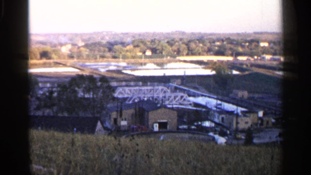 Industrial complex with a white truss structure, open basins, trees, and tall grass in the foreground.