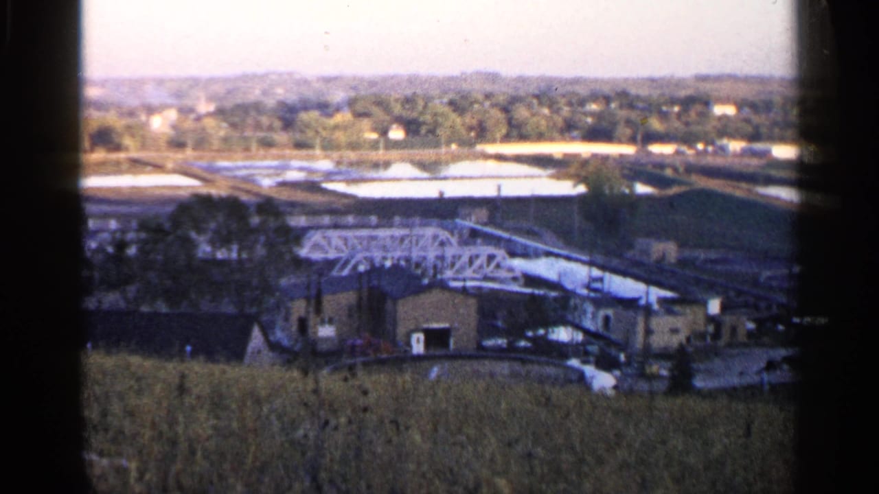 1960 Sioux Falls view of low industrial buildings, a white truss or conveyor structure, open basins, trees, and distant houses.