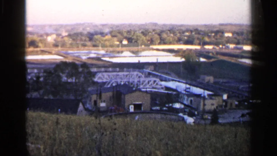 1960 Sioux Falls view of low industrial buildings, a white truss or conveyor structure, open basins, trees, and distant houses.