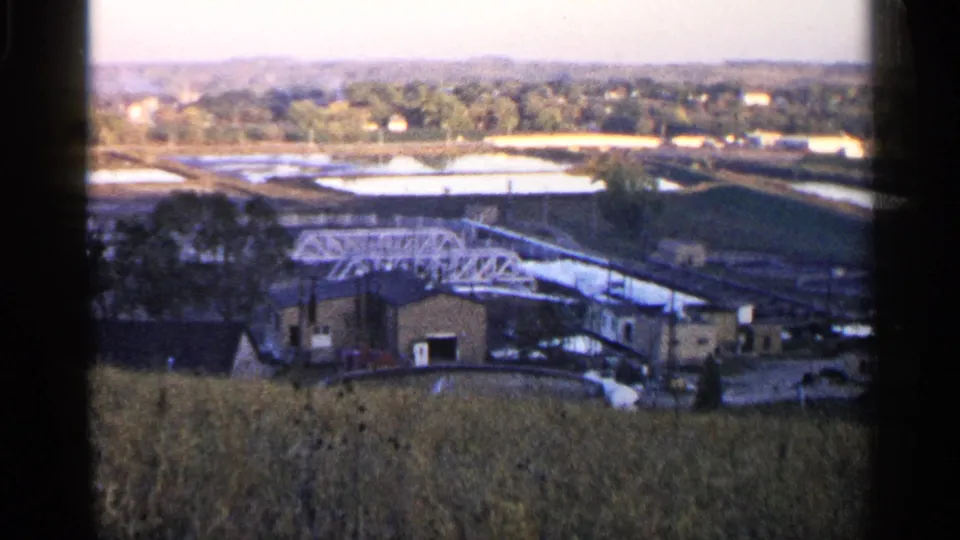 1960 Sioux Falls home-movie frame with open basins above a white truss structure and industrial buildings, viewed past tall grass.