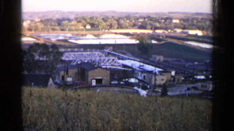 1960 Sioux Falls 8mm frame showing a white truss structure crossing an industrial complex with basins, trees, and distant residential areas.