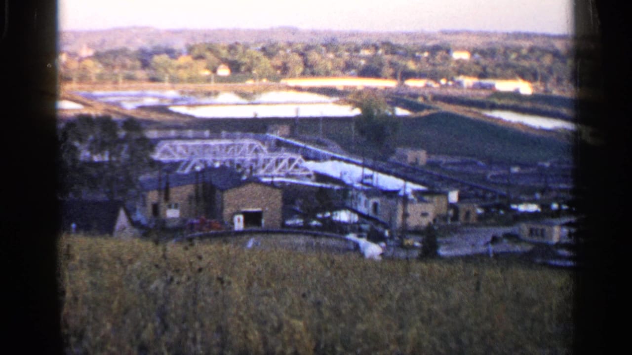 Rural field overlooking low buildings, a white truss bridge, water basins, and distant trees.