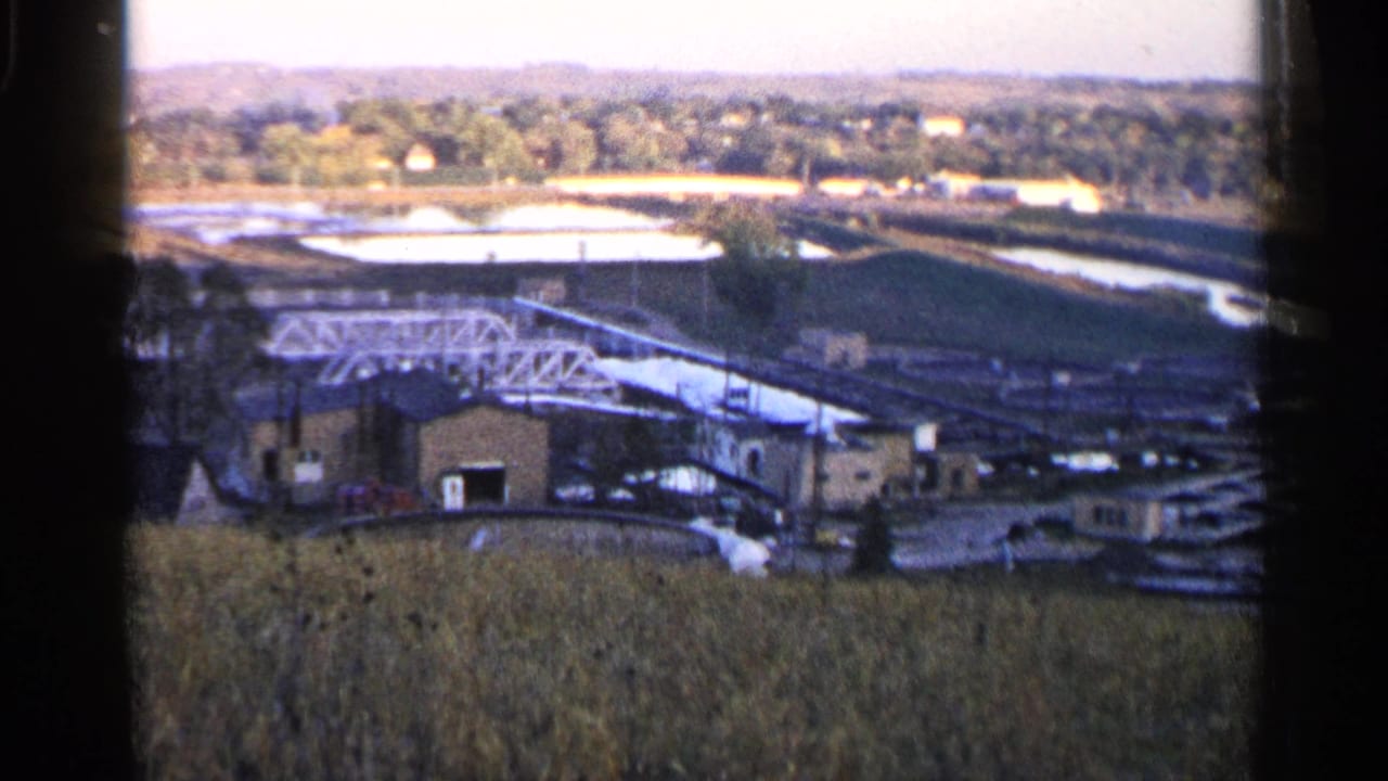 Field and industrial edge with bridge, water basins, river channel, and trees beyond.