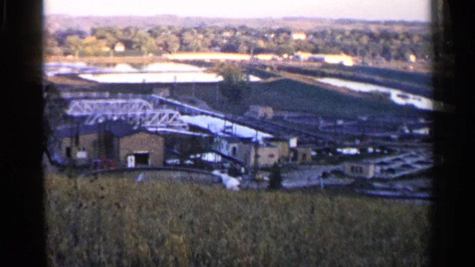 Slightly shifted 1960 Sioux Falls frame with field grasses, low utility-like buildings, a white truss bridge, water basins, and wooded distance.