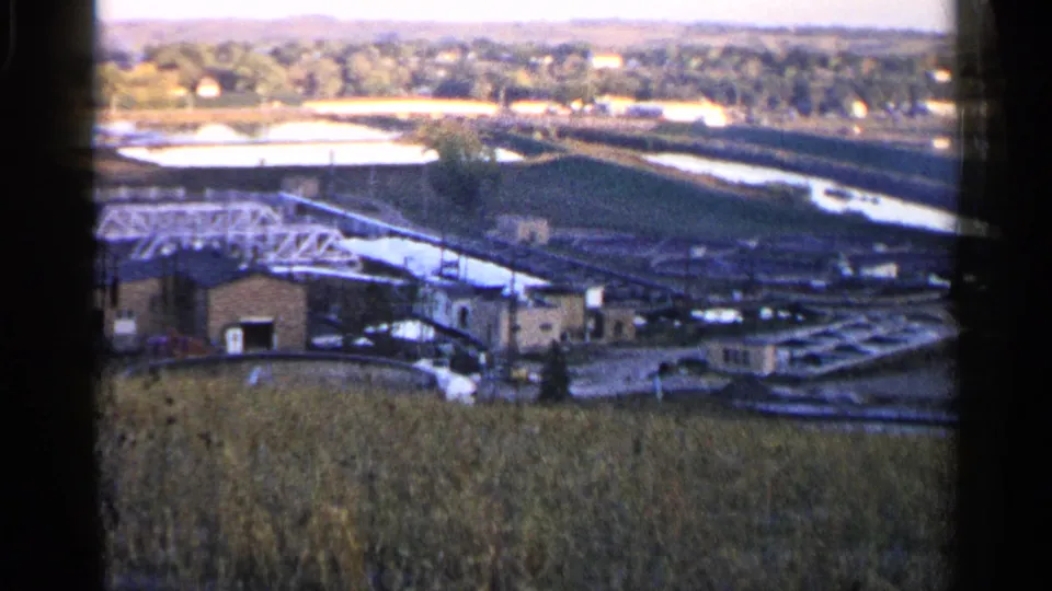1960 Sioux Falls home-movie frame looking over a tan field toward low buildings, a white truss bridge, water channels, bright basins, and trees.
