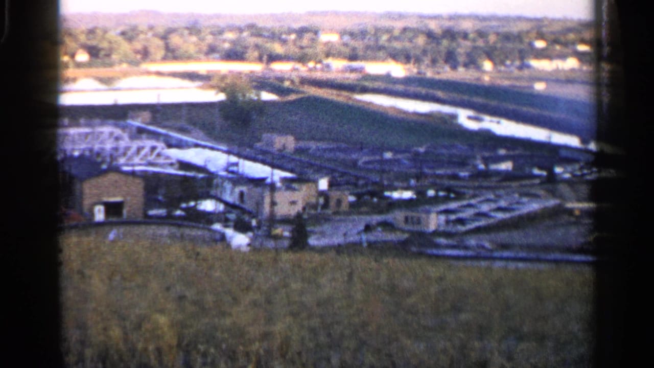 High view across a grassy foreground toward industrial buildings, lines, water, and distant houses.