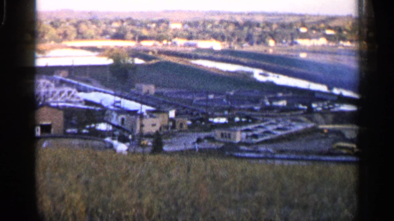 Industrial valley seen from above with a field in front, water channel, buildings, and distant tree-lined houses.