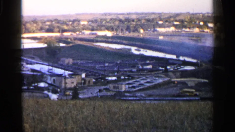 1960 home-movie view near Sioux Falls with soft grain, field foreground, industrial valley, water channel, and distant houses.