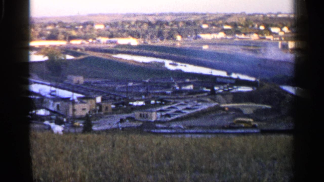 Wide rural view with fields, water channels, distant buildings, and a concrete waterway structure.