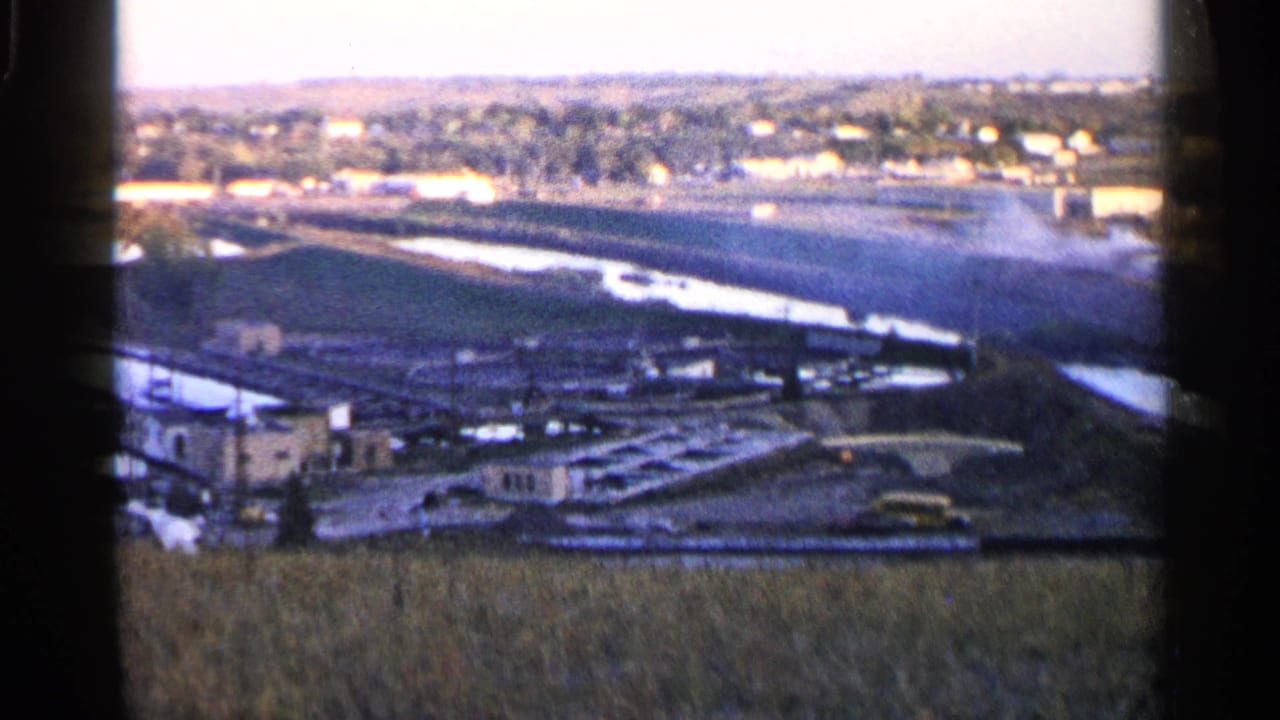 Similar wide view of farmland, water channels, distant buildings, and a small yellow vehicle near the waterway.