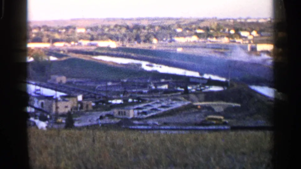 1960 Sioux Falls-area landscape with irrigation or drainage channels, distant settlement, and a yellow vehicle near a concrete waterway.