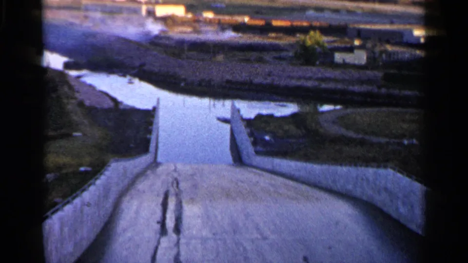 1960 8mm frame looking down a concrete waterway or spillway toward reflective water, bare fields, and distant structures near Sioux Falls.