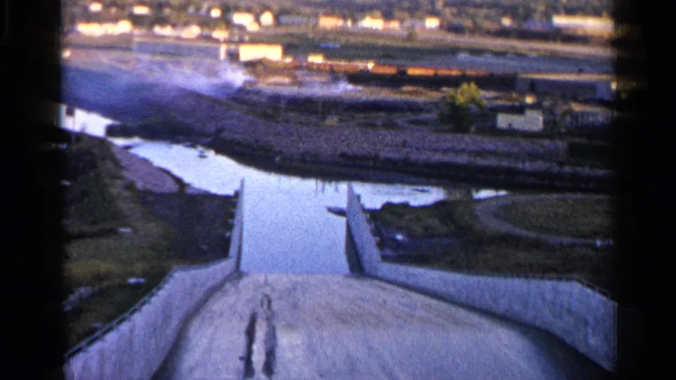 1960 Sioux Falls-area home-movie frame showing a concrete water-control channel descending toward water, fields, and distant buildings.