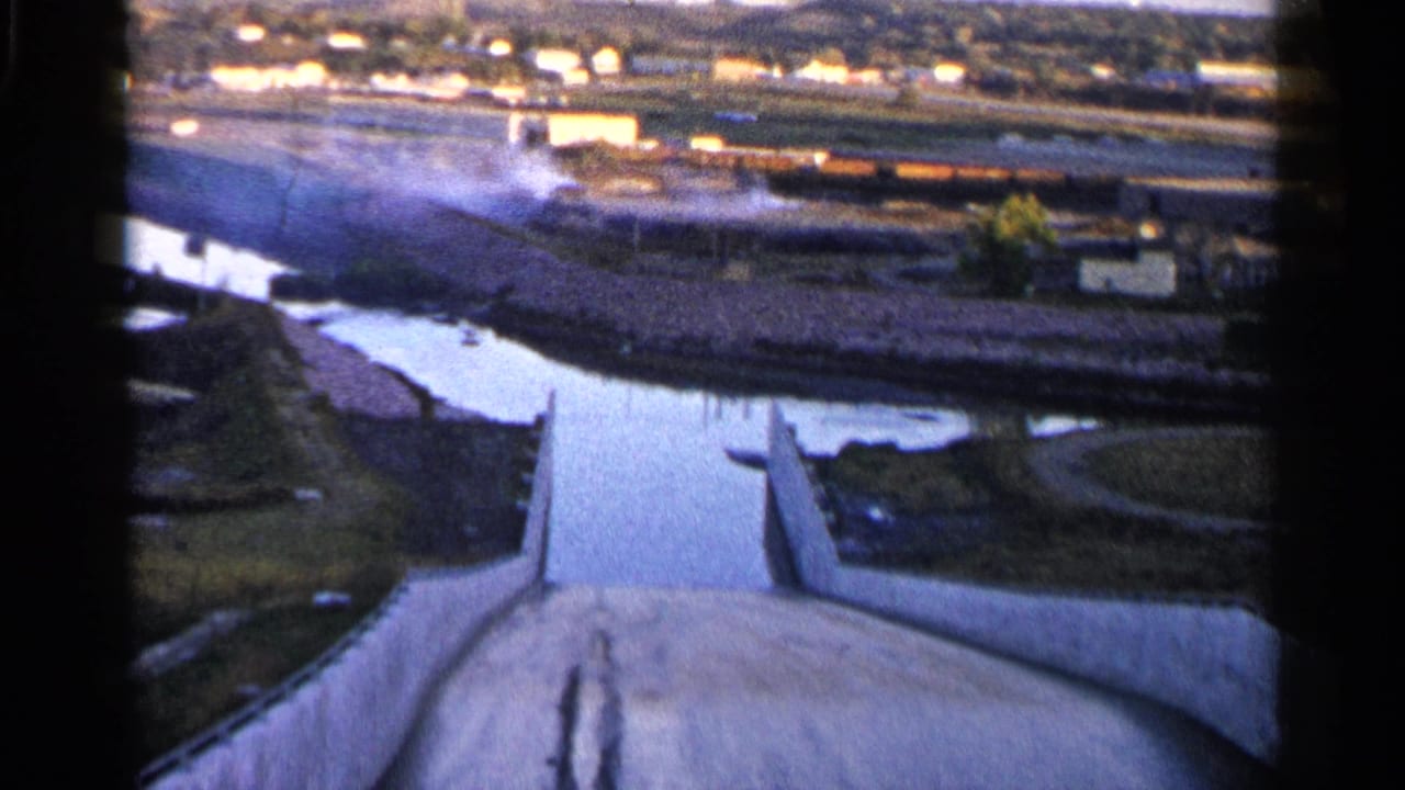 High-angle view of a concrete spillway leading toward a river and distant low buildings.