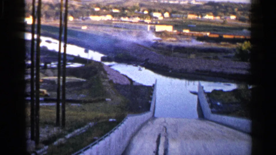 1960 Sioux Falls home-movie frame with a tilted high-angle look over spillway walls, river water, grassy banks, and poles near the channel.