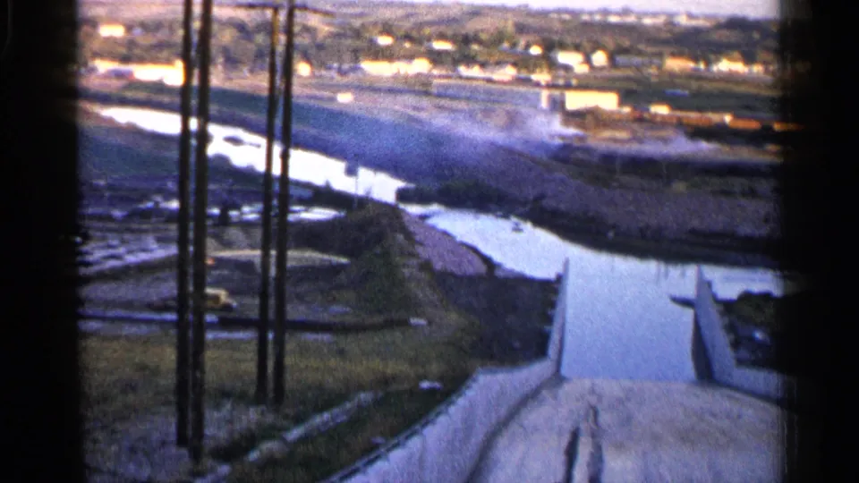 Final 1960 Sioux Falls frame showing the river and concrete spillway from above, with utility poles, grassy ground, and distant low city buildings.
