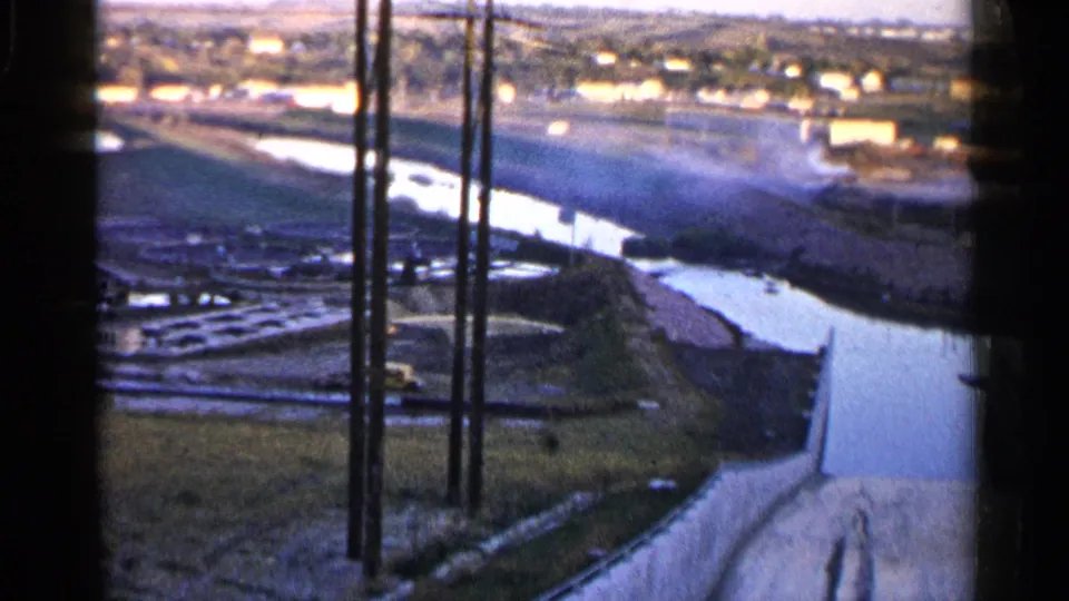 A second 1960 Sioux Falls 8mm frame shows the same channel landscape with utility poles, open land, and a small built-up area beyond.