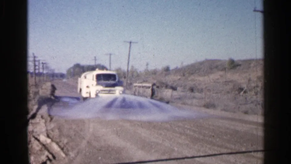 A white truck travels along a rural Sioux Falls road in 1960 as a visible spray or mist spreads near the roadside terrain.