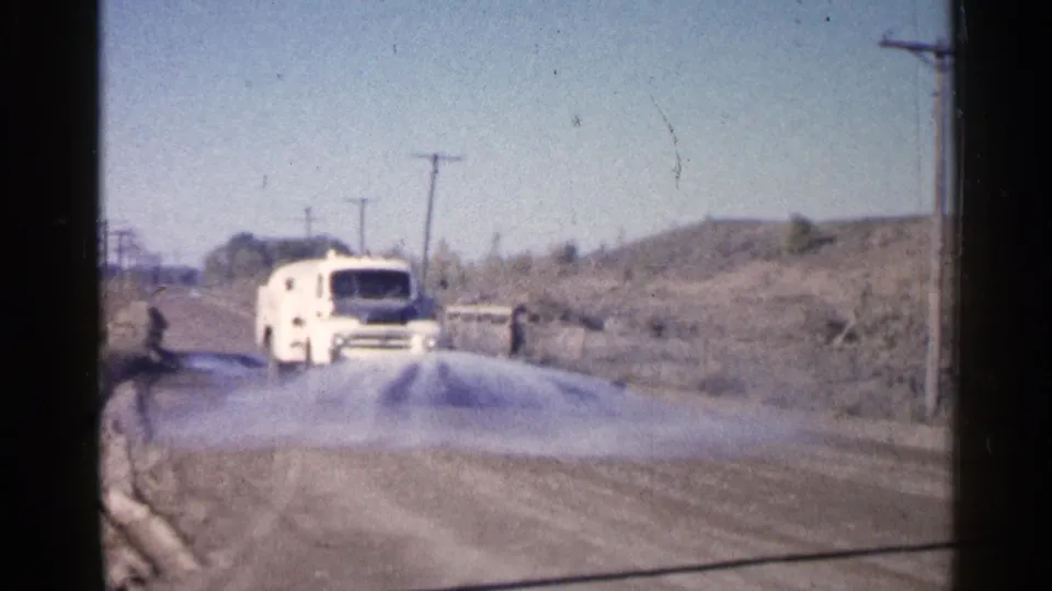 1960 Sioux Falls-area 8mm frame showing a white utility van spraying water on an unpaved roadside under a clear sky.