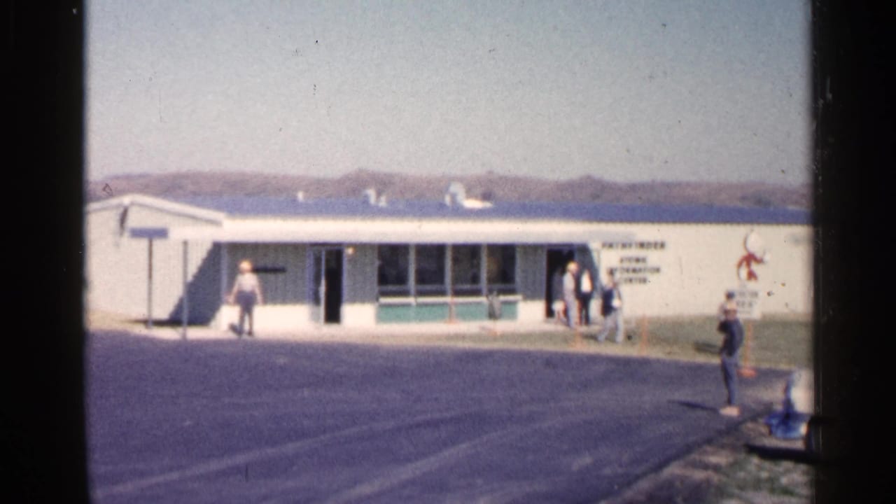 People stand near the entrance of a low building beside a paved lot.