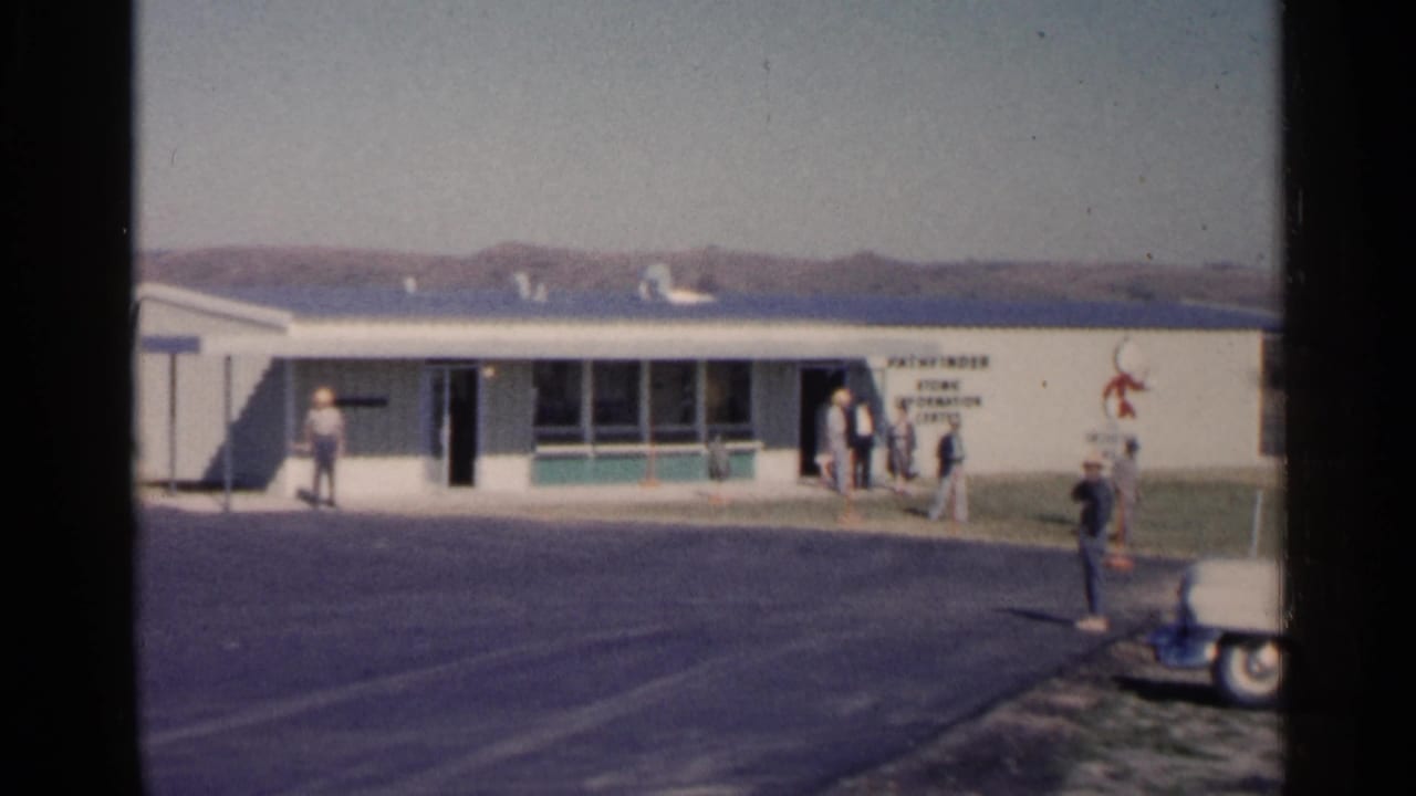 Several people gather by a doorway on a low building with a sign on the wall.