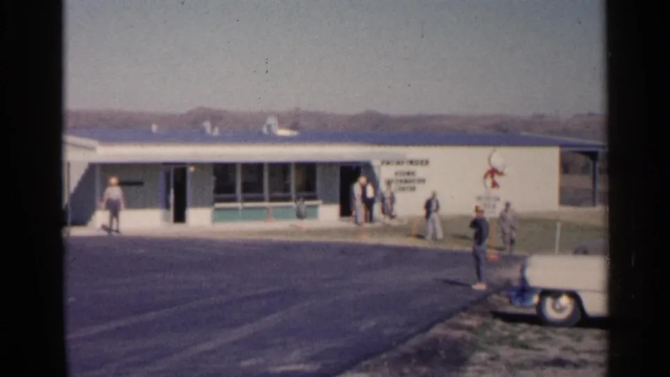 1960 Sioux Falls home-movie frame showing a person in the foreground lot and others near a low building entrance.