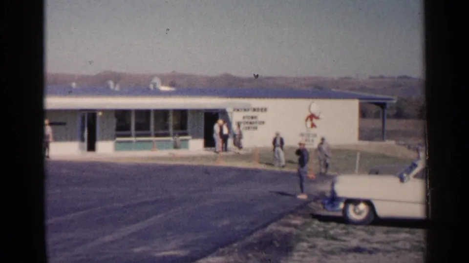 1960 Sioux Falls frame showing a white car at the edge of a paved lot as people stand near a low building entrance.