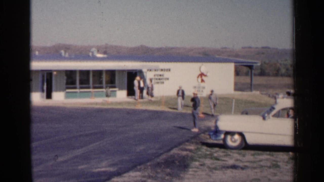 Men near a low building with signage as a white sedan is parked in the foreground.