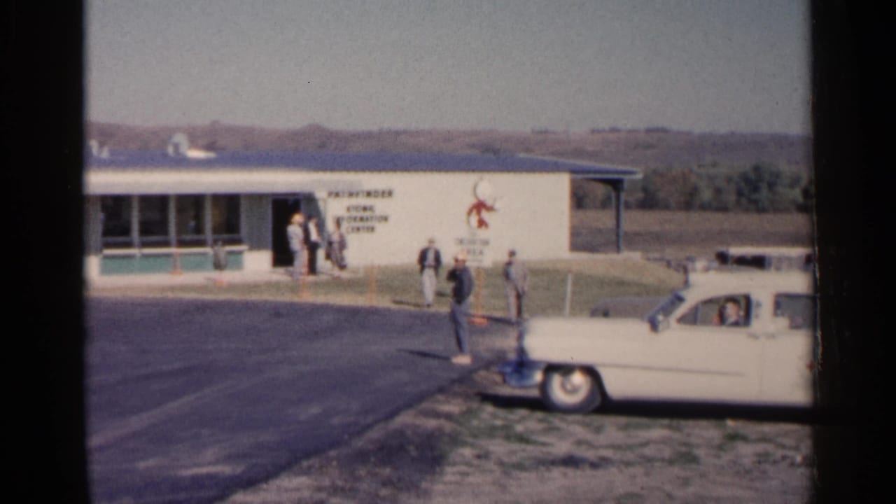 Group of men walk near a signed low building beside a parked white sedan.