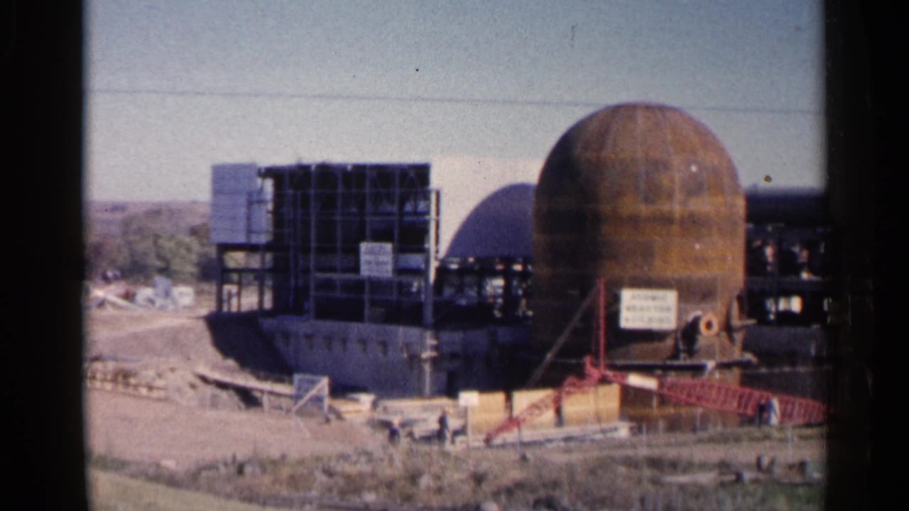 Construction site with spherical vessel, steel framing, red stairs, signage, and workers in the distance.