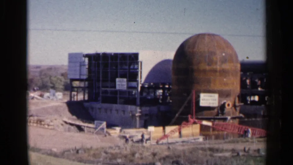 1960 8mm view in Sioux Falls showing a nuclear-facility construction site with a spherical structure, steel framing, signage, and workers.