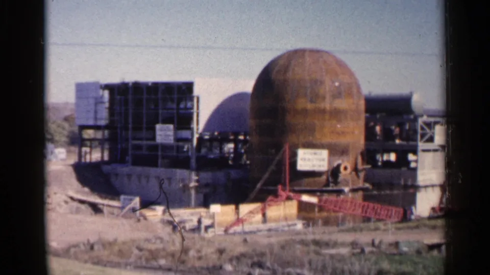 1960 South Dakota home-movie frame showing the nuclear-facility construction area with a spherical vessel, steel frame, and workers.