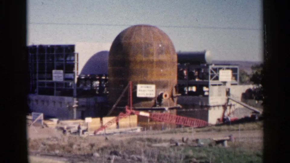 1960 Sioux Falls home-movie frame of a large rusted dome beside exposed framework and a red crane boom at an outdoor construction site.