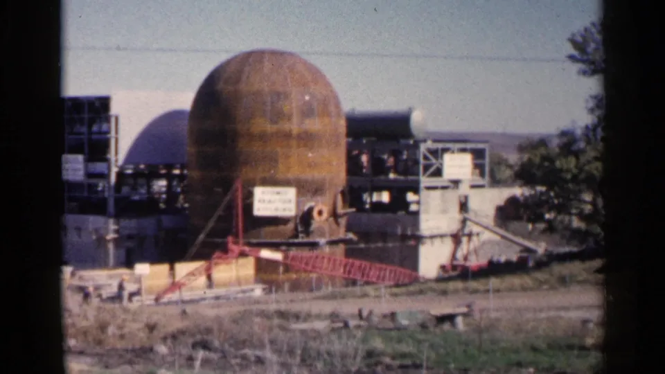 1960 Sioux Falls frame showing the rusted industrial dome, crane arm, scaffolded structures, and trees at the edge of the construction site.